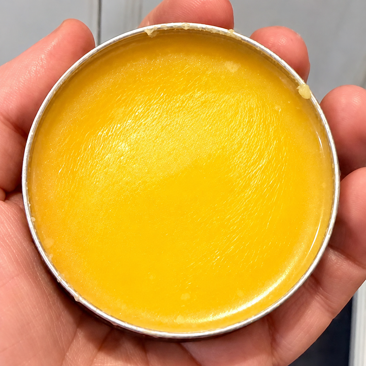 A close-up of a hand holding an open round metal tin filled with bright yellow balm or cream.